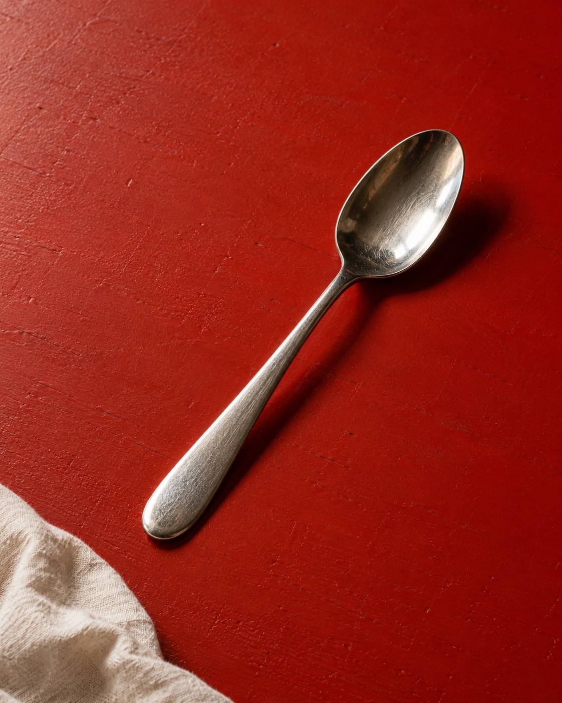An ordinary steel spoon on a red tabletop beside a soft cotton cloth.